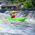 Person kayaking in a green kayak on turbulent water with a house and trees in the background.