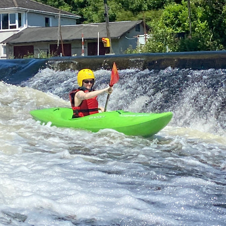 Person kayaking in a green kayak on turbulent water with a house and trees in the background.